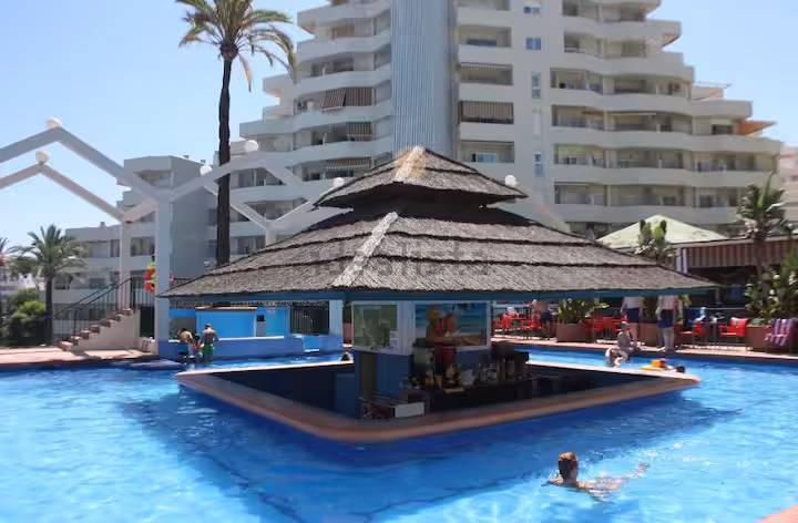 Piscina del complejo con bar de palma y el edificio al fondo